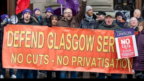 A group of men and women behind a large orange banner that says Defend Glasgow Services - no cuts - no privatisation