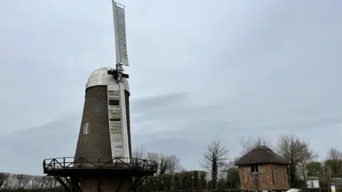 Peter Wilson A bricked windmill with a white top and the two remaining white sails placed in the 'mourning' position. The top sail is just past the 12 o'clock position. The sky in the background is grey with some trees and a small brick house with a thatched roof.