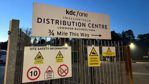 An entryway into the factory. There is a large sign on the gate which says 'KDC/One, swallowfield, distribution centre, lowmoor business park, 3/4 mile this way'. It is getting dark. 