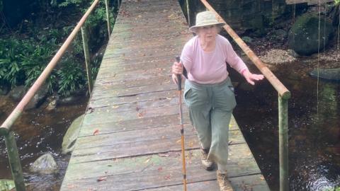 80-year-old Grace Prince in hat, walking boots and hiking trousers, with hiking stick, crossing a bridge over a small river.