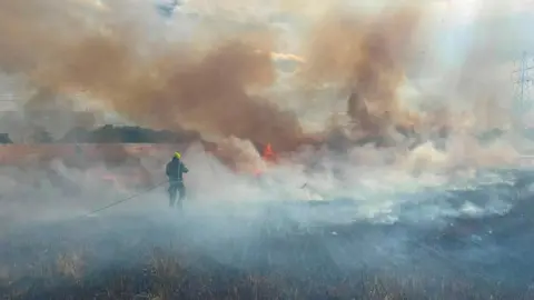 Bedfordshire Fire and Rescue Service A firefighter in full protective gear working to put out a field fire. They are holding a hose and spraying water on to burning vegetation.