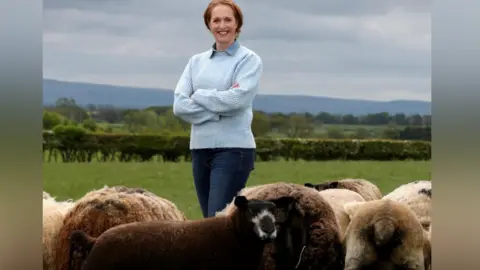 Clare Wise Farmer Clare Wise stands in a field with her arms crossed and is smiling. She has ginger hair is wearing a light blue jumper and jeans. Her flock of sheep are standing in front of her. 