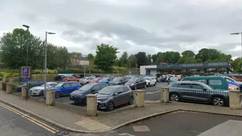 A nearly full car park in front of a single storey Aldi supermarket. Trees in the background.
