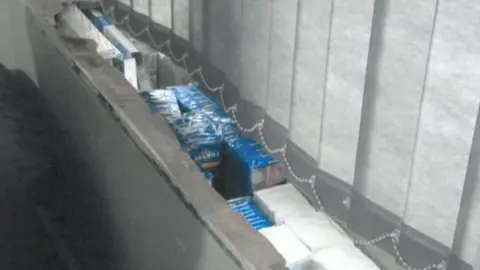 Trading Standards Boxes and packages stacked tightly inside a window ledge behind closed vertical blinds.