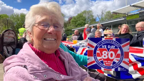 Brenda smiling while holding a VE Day 80th anniversary flag. Behind her are families sitting at Union flag–decorated tables enjoying food, drinks and having conversations. The sky is bright with white clouds and there are green trees in the background. 
