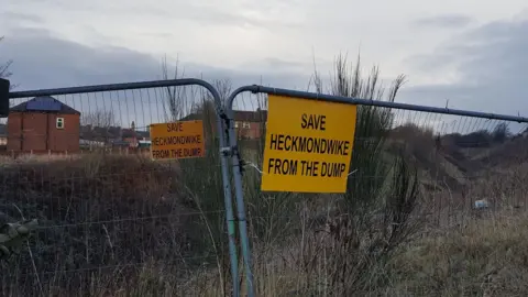 Victoria Harrington Metal fences around a disused area of grass. On them yellow signs read 'Save Heckmondwike from the dump'