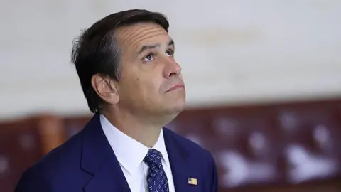A man wearing a blue suit with a white shirt and a blue tie stares up at the ceiling