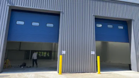 A facility as seen from the outside includes large blue doors that are halfway open. A man is visible inside in a largely empty indoor space.