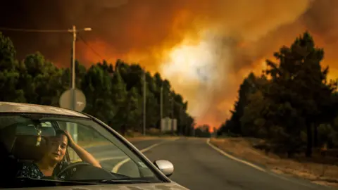 EPA A woman reacts inside her car during a wildfire in Lamosa, Sernancelhe municipality, Viseu district, Portugal, 14 August 2025. She has her left hand on her head and a look of distress on her face, with a fire-coloured sky behind her down the stretch of road 