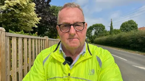 Village resident Stuart Smith stands at the side of the road with his arms crossed looking angry. He is wearing a bright yellow jacket and glasses