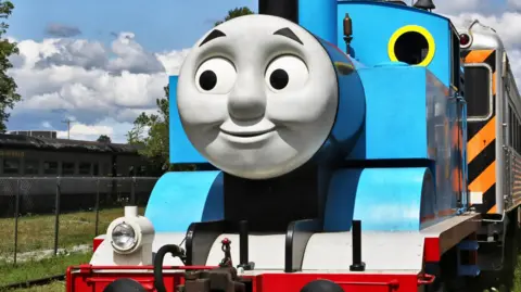 A replica of Thomas the Tank Engine. It is a blue model steam train with a grey smiley face on the front. 