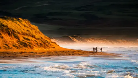 Robert Westerman Golden sunlight illuminates a rugged sand dune on the left, casting warm tones across the windswept beach. Waves roll gently onto the shore, their white foam contrasting with the deep blue water. In the distance, a few figures walk along the shoreline, adding scale.