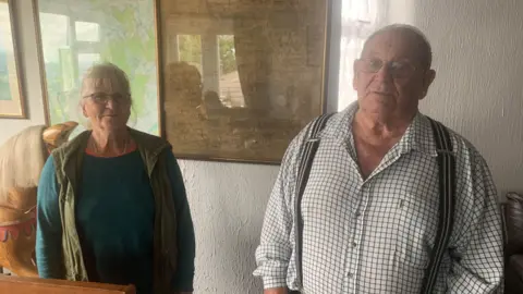 Mick Holder, pictured right, has short hair and is wearing a black and white checked shirt and braces. He is standing next to Forest of Dean Commoners' Association member Sue Adams. Sue has grey hair and is wearing a green long-sleeved shirt under a green gilet.