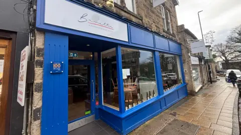 BBC/Jack Conlon The store front of the restaurant Bavette. It is a royal blue colour, with two large windows at the front and a door on the left. Wine bottles and tables and chairs are visible through the windows. There is a white sign with the restaurant name above the door.