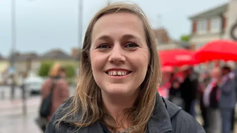 Jen Craft standing in Grays High Street on a rainy day. She is smiling and looking at the camera. Behind her are people with red umbrellas.