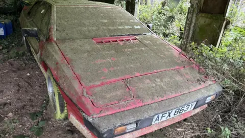 A red Lotus Esprit caked in dirt with moss-covered wheels in an open-sided barn.
