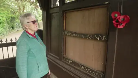 A woman in a green jumper, sunglasses and a priest collar looking at an exposed wooden frame, which is missing its contents.