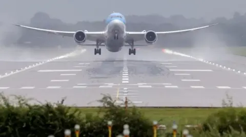 A plane landing at Gatwick Airport. The plane is very close to the ground on the runway, which has water sprayed up each side due to rainy conditions. The plane is white and pale blue. 