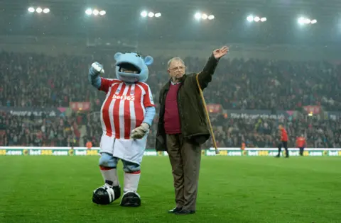 A man is waving to the crowd at Stoke City's stadium and he is standing next to the clubs mascot a blue hippo.