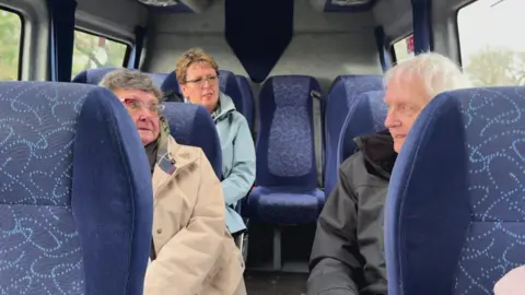 Two women are sitting on the left inside the community bus and a man is sitting on the right side. He is talking to one of the women across the aisle and the lady further back looks like she's listening.
