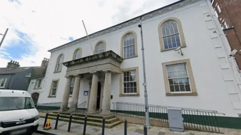 A courthouse with four pillars at the entrance. It's two floors. The windows at the bottom are square and the top windows have oval shapes at the top. It's cloudy. A white van is parked. A cone is on the pavement. 