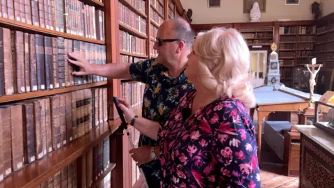 Debbie Ramsey and Daren Smith in the museum at Stonyhurst College. Daren is feeling books on a shelf while Debbie stands next to him.