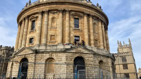 The Radcliffe camera with some protestors, all wearing black, stood on a ledge.