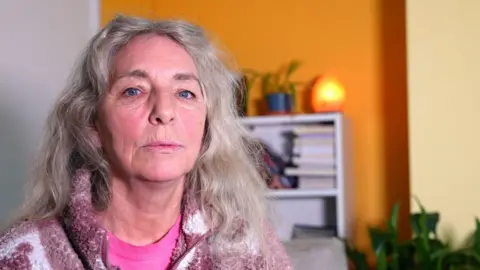 A close, indoor portrait of Janet as an adult with long, wavy, light‑coloured hair. She is wearing a pink top and a patterned fleece. Behind her is a brightly coloured room with a yellow wall, shelving, books, plants, and a softly glowing lamp.