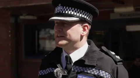 A police chief superintendent, wearing a black police uniform. He is standing outside a police station and is looking side-on from the camera.