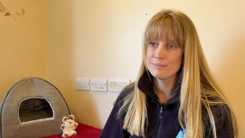 A woman with long blonde hair sits in front of a yellow wall. She is wearing a blue jacket and smiling. There is a pet bed next to her and a small cuddly toy.