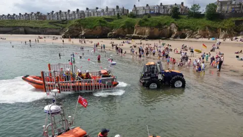 A tractor reverses a trailer carrying a lifeboat into sea at Cullercoats. There are people on the beach but it is not full.
