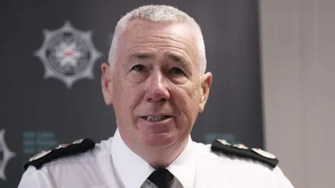 PA Media Chief Constable Jon Boutcher speaking during a press conference at PSNI headquarters, he has white hair and is wearing a white shirt that has black tie and black shoulder pads