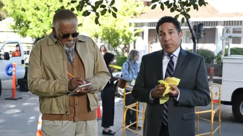 Peacock Oscar Nuñez (right) as his character Oscar Martinez in The Paper, wearing a black suit