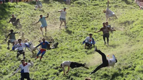 Getty Images A group of men run and tumble down a very steep hill with some of them suspended in mid-air as they fall down. The grassy hill is illuminated by sunlight and dust is being released from the dry ground as they race. Some are in fancy dress, with one dressed as the dog Gromit and another dressed as Superman.