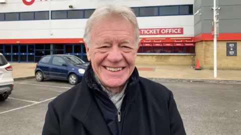 Prof John Fairclough pictured outside Gloucester's stadium. He is wearing a black coat, a black fleece, and a grey scarf, and has grey hair and eyebrows. 