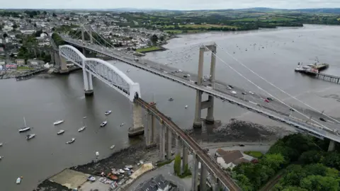 BBC The photo shows an aerial view of the Tamar Bridge, a suspension bridge over the River Tamar. Next to it is a rail crossing. Houses and green fields are seen in the distance. 