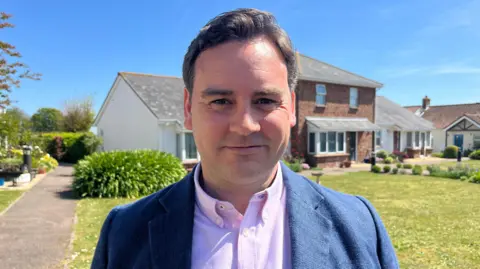 A man with dark brown hair is looking into the camera and smiling. He is wearing a blue blazer over a pink shirt with the top button undone. Behind him there is grass and a path and a row of houses. It's sunny.