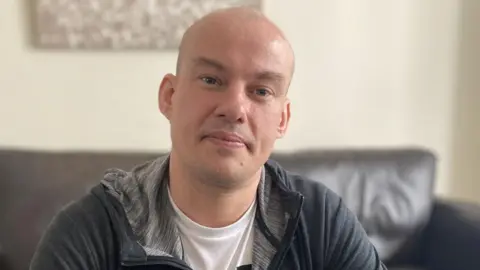 Emma Glasbey/BBC Adam Clarke, a bald-headed man with dark eyes. He is wearing a white t-shirt with a dark zipped top and sat on a black leather sofa.