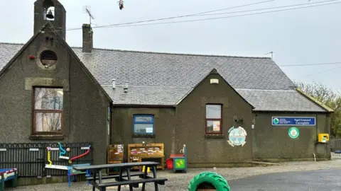 BBC Ysgol Gymuned Carreglefn school - brown grey building with a park bench and children's games on the tarmac