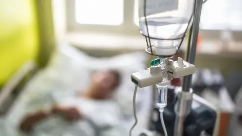 A image of a drip in the foreground in focus with a patient in a hospital bed in the background, out of focus