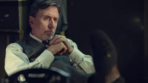 Actor Tim Roth is seated indoors, wearing a light shirt and a buttoned vest, with hands clasped near the chest. The setting appears dimly lit, with muted colours and blurred objects in the background suggesting an older or formal environment.