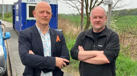 Two men with their arms folded stood a lay-by. A large green field, parking restriction sign and a blue-coloured cafe can all be seen in the background.