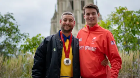 University of Bristol Matt Paine, wearing the Paris silver medal, standing with his arm around Mr Bethell, who is wearing his Team GB red tracksuit top, with the Wills Memorial building out of focus in the background