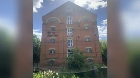 The outside of the former Surrey Advertiser building, which has the name of the paper on the front in black lettering. It is a six-storey mill building in red brick with windows all the way up and greenery growing around the ground floor.