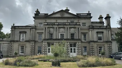 A large mansion-style home is pictured which is the home of Brainkind on a grye cloudy day. A grassed area sits in front of the house which is overgrown in parts. Some vehicles can be seen to the right on the image. 