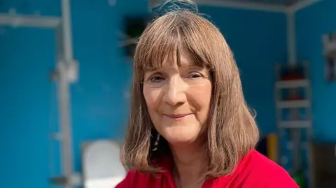 Mary Long-Dhonnau is wearing a red top and smiling at the camera. She has brown hair and is standing in a flood-adapted bathroom, which is in the background but slightly blurred.