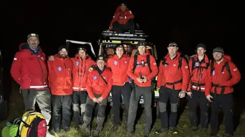 Nine of the rescuers stand in front of a land rover. Another team member is on top of the car. They all wear red jackets and black trousers. There are some backpacks in front of them on the left. It is dark. There are nine men and one woman in the team. 