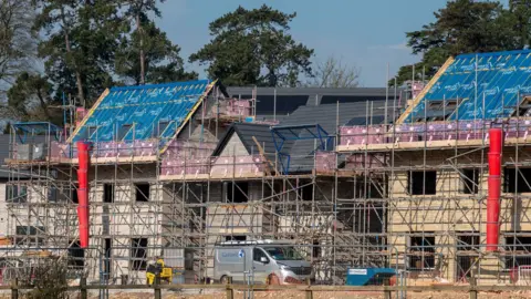 Scaffolding surrounds a couple of new build houses under construction. A silver van is parked outside. Three trees can be seen behind them on what is a sunny day.
