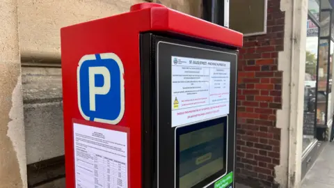 Kris Holland/BBC A close up of one of the electronic parking machines, which is painted red on the sides and top. There is a big blue P sign on the side along with a white laminated sheet showing the charges for each street.