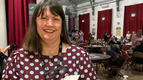 Kerry Hearsey smiles at the camera with tables of people talking behind her. She wears a brown dress with blue polkadots.